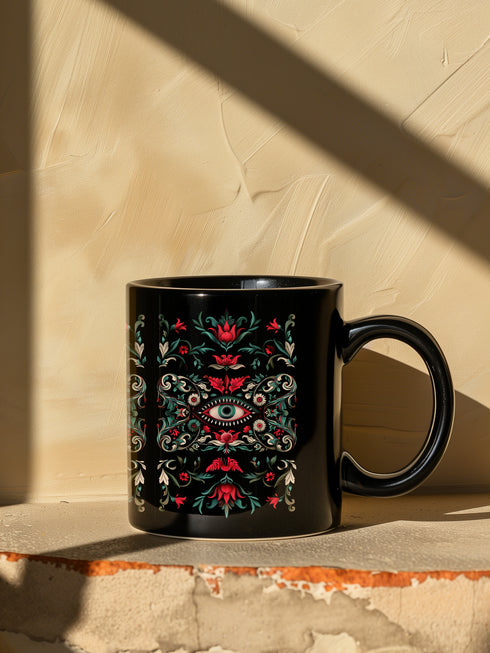 A black ceramic mug featuring a symmetrical folk art design with a central mystic eye motif in turquoise and coral. The design includes ornate scrollwork, red tulip-like flowers, sage green leaves, and cream flourishes arranged in a decorative pattern. The mug has a glossy finish with a C-shaped handle.